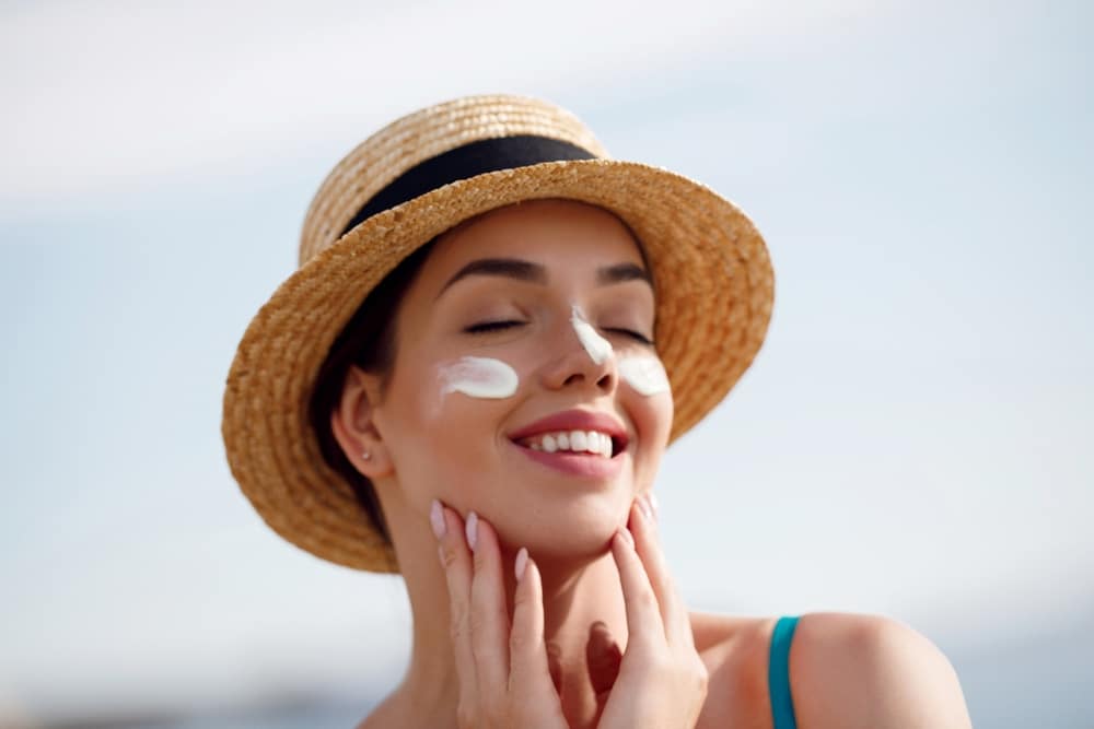 Smiling woman in sun hat applying sunscreen on cheeks, emphasizing sun protection to prolong Botox benefits.