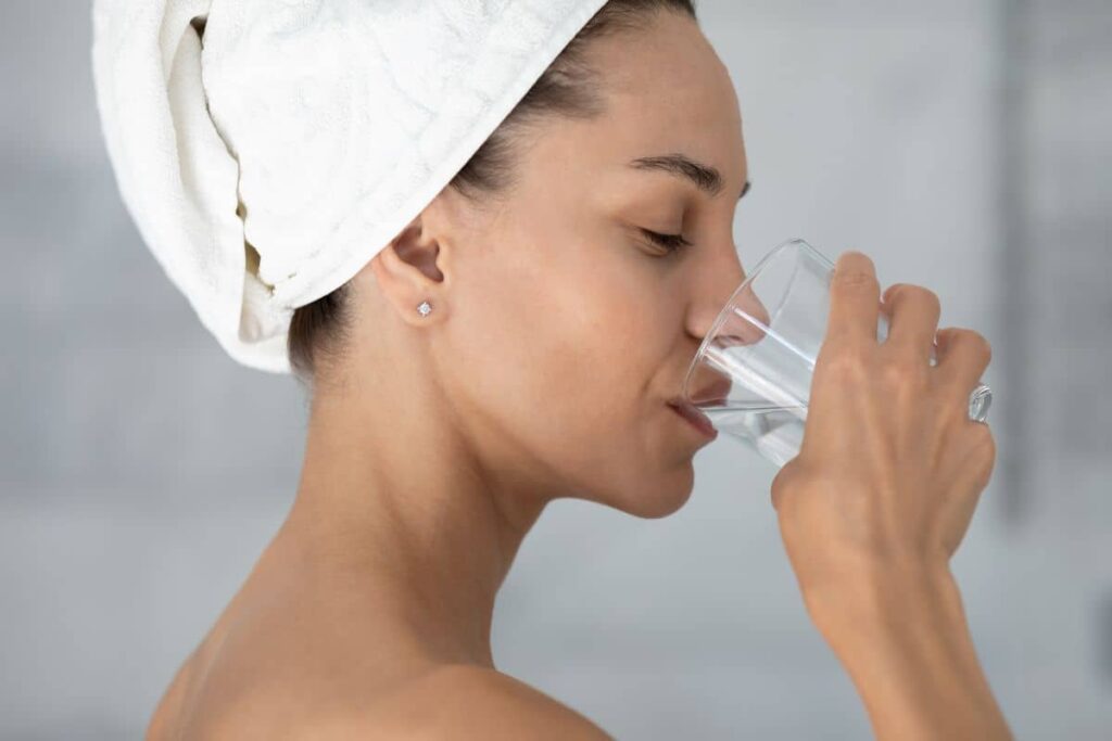 Woman drinking water with towel-wrapped hair, representing hydration as part of rhinoplasty pre-op preparation.