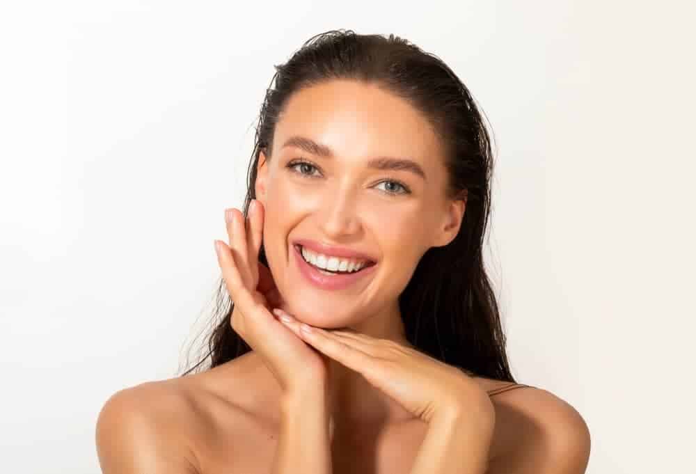 A smiling woman with wet-look dark hair rests her chin on her hands, showcasing a smooth complexion and bright smile against a white background.