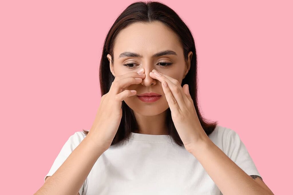A woman with dark hair against a pink background looking down and gently touching both sides of her nose to examine the skin and tip.